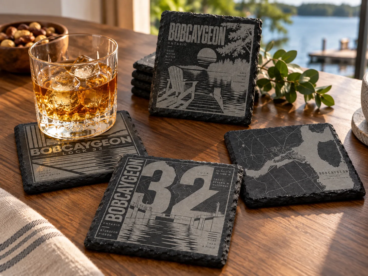 Engraved slate coasters with Bobcaygeon designs on a table by a lake, next to a whiskey glass.
